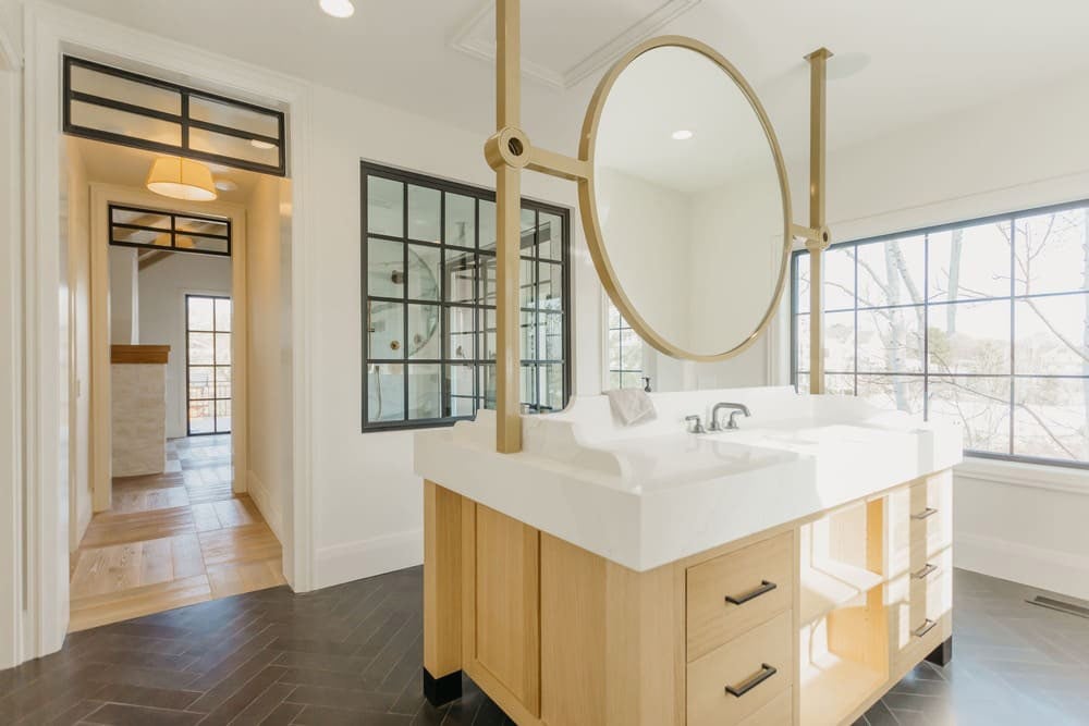 A modern bathroom featuring a double vanity with wooden cabinetry, a large circular mirror, black-framed frosted glass doors, and herringbone tile flooring.